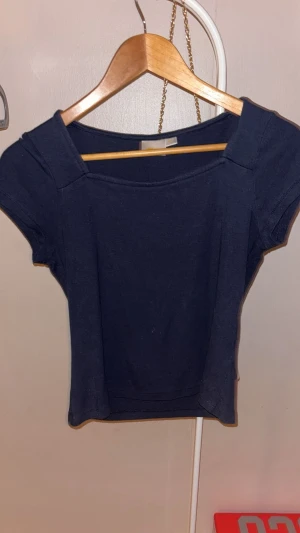 Mörkblå croppad topp med kort ärm - En stilren mörkblå croppad topp med fyrkantig ringning och korta ärmar. Toppen har en enkel design och är tillverkad i mjuk bomull, perfekt för en avslappnad look. Passformen är normal och toppen är lätt att matcha med olika outfits.