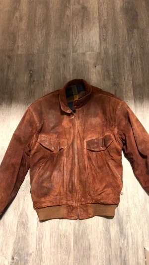 Brun mockajacka med dragkedja - Snygg brun mockajacka med dragkedja framtill, två bröstfickor med lock och knappar samt ribbade muddar i midjan. Jackan har en klassisk bomberstil och är fodrad med rutigt tyg på insidan av kragen. Perfekt för dig som gillar vintagevibbar.