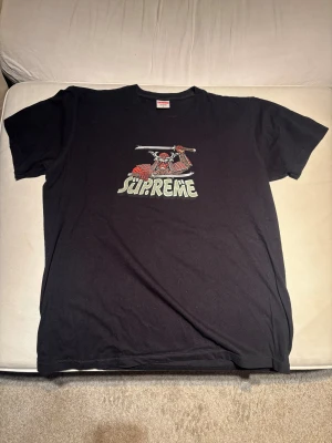 Svart Supreme t-shirt med samurai-tryck - Svart t-shirt från Supreme med coolt samurai-tryck och Supreme-logga på bröstet. Klassisk passform med rund hals och korta ärmar. Tillverkad i mjuk bomull som är skön att bära. Perfekt för dig som gillar streetwear och unika prints.