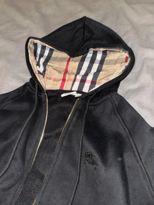 Svart Burberry hoodiejacka med dragkedja - Svart hoodiejacka från Burberry med klassiskt rutigt mönster i huvan. Jackan har guldfärgade detaljer på dragkedjan och snörning, samt diskret Burberry-logga broderad på bröstet. Perfekt för dig som vill ha en stilren och lyxig look.