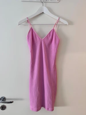Rosa ribbad kortklänning. chiquelle - Supersöt rosa ribbad klänning med smala axelband och v-ringad hals. Klänningen är figurnära och kort, perfekt för varma dagar. Materialet är stretchigt och mjukt, vilket gör den riktigt bekväm att bära.