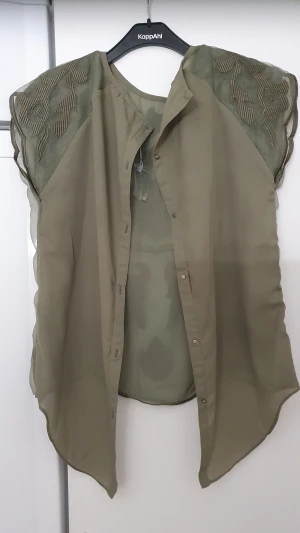 Blus - Snygg olivgrön blus i transparent tyg från Oxygene. Blusen har korta ärmar och är dekorerad med broderade blommor och blad på axlar och rygg. Knäpps framtill med tryckknappar och har en asymmetrisk nederkant. Perfekt för dig som gillar unika detaljer. Finns bara hål annars är den i bra skick mycket bra skick.