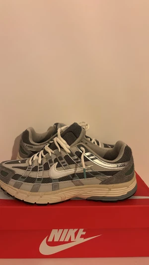 Nike P-6000 grå sneakers - Nike P-6000 sneakers i grå och silver med mesh och syntetpaneler, vita snören och retro löparstil. Snygga detaljer med broderad Swoosh och texten P-6000 på sidan. Platt sula och rund tå för skön komfort. Perfekt för dig som gillar sportig vibe.