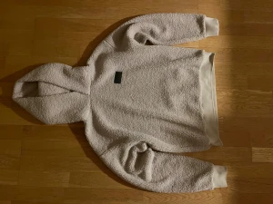 Beige fleece hoodie Scotch & Soda - Mysig beige hoodie från Scotch & Soda i storlek S. Tröjan är tillverkad i mjuk fleece med huva och ribbade muddar vid ärmslut och nederkant. En diskret grön patch med logga på bröstet ger en clean look. Perfekt för kyliga dagar. Har används ett fåtal gånger 