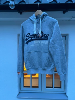 Grå Superdry hoodie med tryck - Snygg grå hoodie från Superdry med stort svart och vitt tryck på bröstet. Klassisk känguruficka framtill, ribbade muddar och vit dragsko med metalländar. Mjuk bomullsblandning och orange Superdry-detalj på snöret. Perfekt för chill och streetwear.
