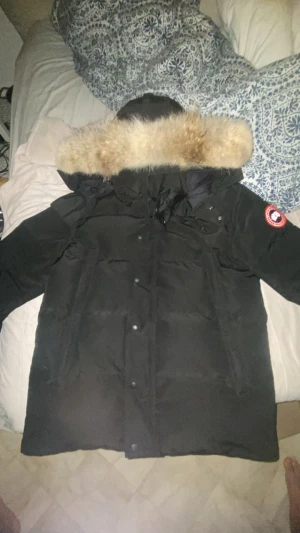 Svart Canada Goose dunjacka med päls - Säljer en svart dunjacka från Canada Goose med ikonisk röd logga på ärmen och stor fluffig päls runt huvan. Jackan har flera fickor, dragkedja och knappar framtill. Perfekt för kalla vinterdagar och har en riktigt schysst oversized look.