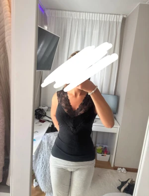 Svart linnetopp med spetsdetaljer - Snygg svart linnetopp med djup v-ringning och breda spetskanter längs halsringningen. Toppen är ärmlös och har en figurnära passform som framhäver siluetten. Perfekt att styla med jeans eller kjol för en trendig look.