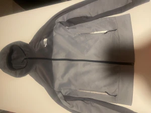 Grå/Blå Full zip Hoodie från The North Face  - Snygg grå/Blå Full Zip från The North Face med huva och hel dragkedja. Hoodien har två fickor med dragkedja och reflekterande detaljer. Perfekt för dig som gillar streetwear och vill ha en clean look. Logga på bröstet och sportig passform.