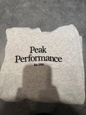 Grå Peak Performance hoodie - Snygg ljusgrå hoodie från Peak Performance med broderad logga på bröstet. Tillverkad i bomull och polyester. Storlek 170 men sitter som en S.