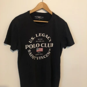 Svart t-shirt Polo Club Saint Vincent - Svart t-shirt från Polo Club Saint Vincent med rund hals och vit textprint framtill. Trycket har en amerikansk flagga och texten 'US. Legacy Made in the US. Polo Club Saint Vincent'. Skön passform och klassisk stil, perfekt till jeans.