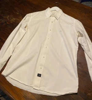 Vit skjorta från Legacy, storlek M - Klassisk vit skjorta från Legacy i storlek M. Skjortan har en stilren design med krage, knappar hela vägen och diskret broderad logga på bröstet. Tillverkad i mjuk bomull som känns skön mot huden. Perfekt för dig som gillar enkel och clean stil.