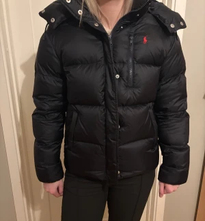 Svart/ marinblå pufferjacka från Polo Ralph Lauren - Svart pufferjacka från Polo Ralph Lauren med röd logga på bröstet. Jackan har huva, dragkedja och knappar framtill samt flera fickor. Den är vadderad och har en klassisk, lite boxig passform som är perfekt för kalla dagar. Det ända som inte är som nytt på jackan är den flärpen som man hänger jackan i, den har gått sönder lite (går att se på sista bilden)