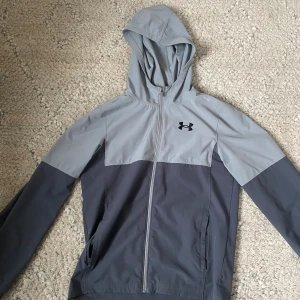 Grå vindjacka från Under Armour - Snygg tvåtonad grå vindjacka från Under Armour med huva och hel dragkedja. Jackan har elastiska muddar med logotypdetalj, sidofickor och långärmad design. Perfekt för träning eller chill dagar ute. Lätt och smidig polyester som andas.