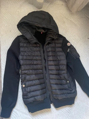 Svart Moncler pufferjacka med huva - Svart pufferjacka från Moncler med stickade ärmar och quiltad front. Jackan har en stor huva, dragkedja framtill och två fickor med tryckknappar. Moncler-logga på ärmen och en extra ficka med dragkedja på vänster arm. Perfekt för kyliga dagar.