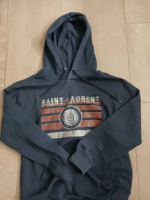 Snygg mörkblå hoodie från Saint Laurent med tryck framtill i rött, vitt och blått. Tröjan har huva med snörning och en klassisk känguruficka. Perfekt för dig som gillar streetstyle och vill ha något bekvämt men ändå stilrent. En perfekt julklapp till dig som är intresserad av kläder. skriv vid funderingar, priset är ej hugget i sten. 