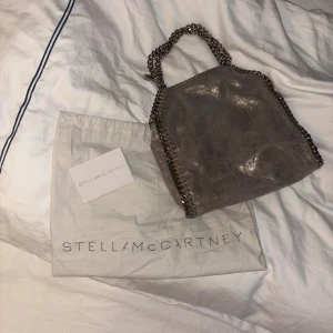 Grå axelväska Stella McCartney - Snygg grå axelväska från Stella McCartney med glittrig yta och kedjedetaljer längs kanterna och handtaget. Väskan är i nyskick använd endast fåtal gånger kommer med kvitto , dustbag och lappar 