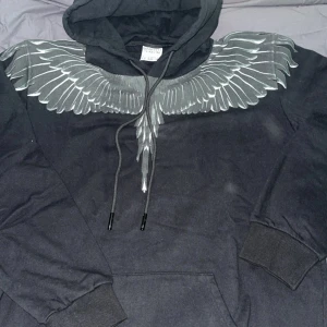 Svart hoodie Marcelo Burlon med vingar - Svart hoodie från Marcelo Burlon County of Milan med ikoniskt vingtryck över axlar och bröst. Tröjan har huva med snörning, känguruficka och ribbade muddar. Grym streetwear-vibe och perfekt för dig som gillar unika detaljer. Priset kan diskuteras. Vid frågor så är det bara att skriva. Är 178, 60kg och den är lite för stor på mig 