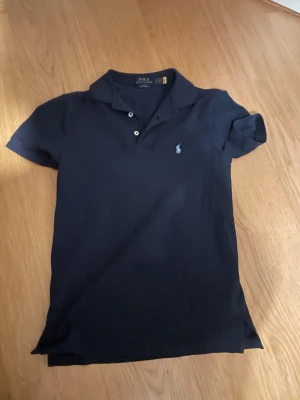 Mörkblå pikétröja från Polo Ralph Lauren - Säljer en mörkblå pikétröja från Polo Ralph Lauren i slim fit med stretch mesh. Tröjan har klassisk krage, korta ärmar och tre knappar framtill. Den ikoniska logon är broderad på bröstet. Perfekt för dig som gillar stilrena och tidlösa plagg.