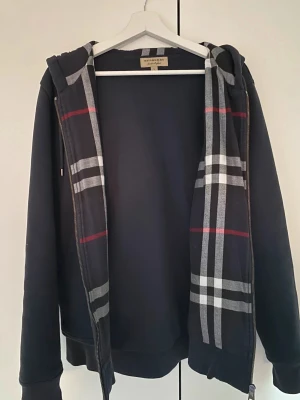 Svart Burberry zip hoodie  - Snygg svart hoodie från Burberry med klassiskt rutmönster i grått, vitt och rött på insidan av huvan och längs dragkedjan. Tröjan har huva, långa ärmar och ribbade muddar. Perfekt för dig som gillar stilrena streetwear-plagg med lyxig touch.