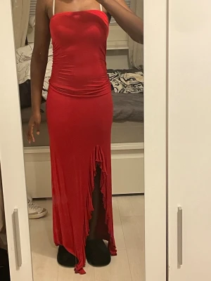 Röd långklänning med slits - Säljer en röd långklänning med tight passform och snygg rynkdetalj längs sidorna. Klänningen är axelbandslös och har en hög slits framtill med volangkant. Materialet är stretchigt och följsamt, perfekt för dig som vill sticka ut. Använd bara en gång. Storlek s.