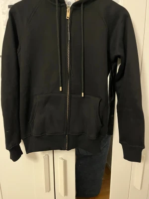 Svart Burberry hoodie med dragkedja - Svart hoodie från Burberry med klassisk rutmönstrad huva, guldfärgade detaljer på snöret och dragkedja framtill. Mjuk bomullsmix, två fickor fram och ribbade muddar. Snyggt Burberry-tryck på snörändarna och insidan av huvan.