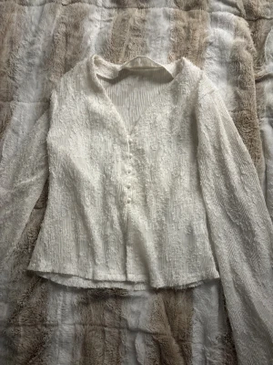 Vit blus med struktur och knappar - Supersöt vit blus med långärmade transparenta ärmar och v-ringning. Blusen har en unik strukturerad yta och fina tygklädda knappar framtill. Passformen är normal och materialet känns lätt och luftigt, perfekt för vår och sommar.