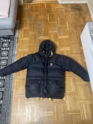 Svart dunjacka från Fjällräven S - Svart dunjacka från Fjällräven med huva och gula snören. Jackan har två stora fickor framtill med knappar och dragkedja. Klassisk Fjällräven-logga på bröstet och detaljer i silver. Perfekt för kalla dagar och riktigt snygg streetstil.