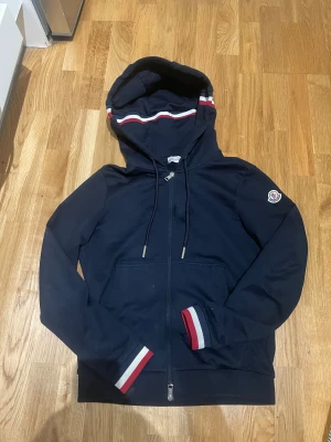 Mörkblå Moncler hoodie med dragkedja - Har nfc skan. Snygg mörkblå hoodie från Moncler med dragkedja och huva. Röd och vit rand på huvan samt muddar med samma färger. Klassisk Moncler-logga på vänster ärm. Mjuk bomullskänsla och fickor framtill. Perfekt för dig som gillar stilrena streetwear-plagg.