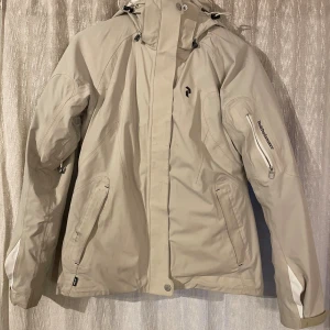 Peak Performance beige vindjacka S - Peak Performance skidjacka i beige med avtagbar svart innerjacka. Jackan har huva, dragkedja och flera fickor, samt Peak Performance-logga på bröstet och ärmen. Perfekt för kalla dagar. Snygg, funktionell och stilren design. Den har några fläckar och ett litet litet hål i kragen. Skicka om bilder på detta önskas💕