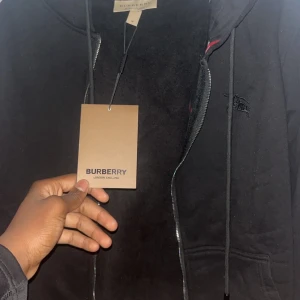 Svart hoodie från Burberry med dragkedja - Svart hoodie från Burberry med klassisk logga broderad på bröstet och dragkedja framtill. Insidan har det ikoniska rutiga Burberry-mönstret i svart, vitt och rött. Tröjan har huva med snörning och känguruficka. Perfekt för en stilren och lyxig look.