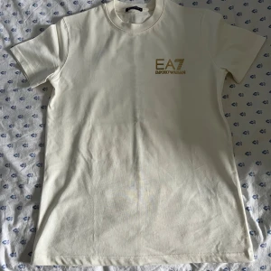 Vit EA7 t-shirt - Snygg vit t-shirt från EA7 Emporio Armani med guldigt tryck på bröstet och längs ryggen. Klassisk rund hals och korta ärmar. Tillverkad i mjuk bomull som känns skön mot huden. Perfekt för dig som gillar stilrena och exklusiva plagg. Helt ny, Pris kan diskuteras vid snabb affär.