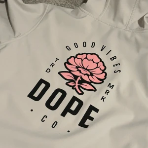 Skidjacka från DOPE - Dope med huva och snörning. Framtill och på ryggen finns ett stort tryck med svart text och en rosa blomma. Tröjan har känguruficka och ribbade muddar. Perfekt för dig som gillar streetwear och vill sticka ut med coolt tryc