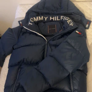 Blå pufferjacka Tommy Hilfiger - Marinblå pufferjacka från Tommy Hilfiger med huva och dragkedja. 
