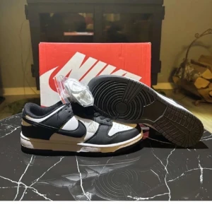 Nike Dunk Low svart/vit sneakers - Säljer ett par Nike Dunk Low sneakers i klassisk svart och vit färg. Skorna har en låg silhuett, svart swoosh och snörning. Ovandelen är i skinn med perforerad tåbox för extra andningsförmåga. Perfekta för dig som gillar streetstyle och vill ha en tidlös look.
