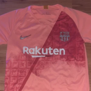 Barcelona Messi 10 rosa fotbollströja Nike - Säljer en officiell FC Barcelona fotbollströja från Nike i rosa med tryck av Messi 10 på ryggen. Tröjan har korta ärmar, V-ringning och klubbmärke samt Rakuten-logga på bröstet. Materialet är lätt och andas, perfekt för match eller träning. (Obs den är rätt använd och lite av trycket där bak går lite av kolla sista bild)