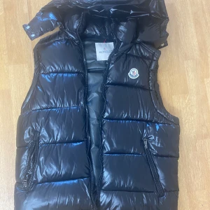 Svart dunväst från Moncler - Snygg svart dunväst från Moncler med glansig finish och klassisk logotyp på bröstet. Västen har quiltad design, två dragkedjeförsedda fickor och hög krage. Perfekt för lager-på-lager och streetstyle. Stängs med dragkedja framtill.