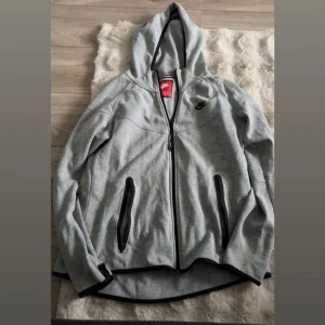 Grå hoodie från Nike med dragkedja - Snygg grå hoodie från Nike med hel dragkedja och svarta detaljer. Tröjan har huva, två fickor med dragkedja och svart Nike-logga på bröstet. Perfekt för chill eller träning. Stl är XL men känns mer som en M-L