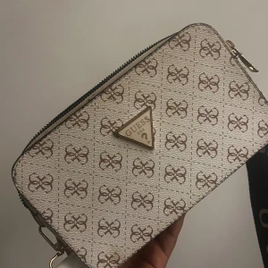  axelväska från Guess med logomönster -  axelväska från Guess med logomönster och justerbar svart axelrem med Guess-tryck. 