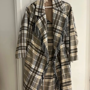 Rutig kappa med fransar från H&M XS - Snygg rutig kappa från H&M i beige, grått och brunt med stora slag och fransar längs kanterna. Oversized känsla och mjukt ull-liknande material. Perfekt för dig som vill sticka ut med en trendig och mysig look.