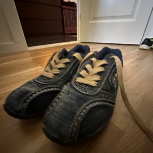 Diesel denim sneakers med beige snörning - Unika sneakers från Diesel i mörkblå denim med kontrastsömmar och beige snörning. Skorna har rund tå, platt sula och coola detaljer som nitar och broderad logga på sidan och hälen. Perfekta för dig som vill sticka ut med en streetinspirerad look.