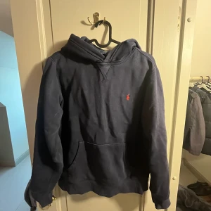 Mörkblå hoodie från Polo Ralph Lauren - Mörkblå hoodie från Polo Ralph Lauren med klassisk huva och känguruficka. Tröjan har det ikoniska röda Polo-logot på bröstet och är tillverkad i mjuk bomull. Perfekt för dig som gillar en avslappnad och stilren look. Tröjan är i storlek barn L14-16.