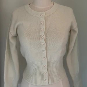 Beige stickad kofta med knappar - Snygg beige stickad kofta med rund hals och långa ärmar. Koftan har ribbad nederkant och muddar samt en rad med matchande knappar framtill. Perfekt för lager-på-lager och enkel att styla till olika outfits.