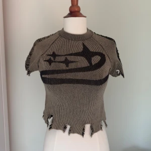 Stickad croppad topp med stjärnmönster - Unik croppad stickad topp i hår/beige och svart med råa kanter och ett stort stjärnmönster framtill. Toppen har korta ärmar och coola slitna detaljer längs fållen och axlarna. Perfekt för dig som vill sticka ut med en edgy stil.
