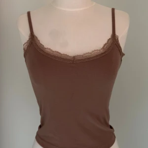 Brun linnetopp med spetskant - Säljer en brun linnetopp med smala axelband och fin spetskant längs urringningen. Toppen är figurnära och tillverkad i mjukt bomullsmaterial, perfekt för en avslappnad och trendig look.