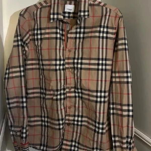 Burberry rutig skjorta i bomull - Klassisk rutig skjorta från Burberry i beige, svart, vitt och rött. Skjortan har lång ärm, knappar framtill och ikoniskt rutmönster. Tillverkad i mjuk bomull och har en normal passform. Perfekt för dig som gillar tidlös och trendig stil.super stor i storlek 