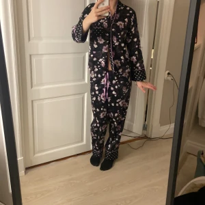 Svarta blommiga pyjamasbyxor med prickiga detaljer med matchande tröja  - Säljer ett par svarta pyjamasbyxor med lila och vita blommor, samt prickiga detaljer vid benslut och midja samt tröja gjort av silk . Byxorna har en avslappnad passform och snörning i midjan med lila band. Materialet är mjukt och glansigt, perfekt för chillkvällar.