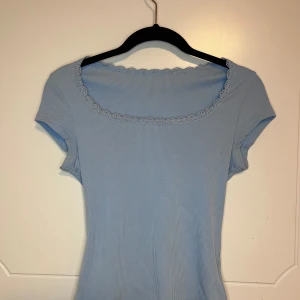 Ljusblå ribbad topp med spetskant - Supersöt ljusblå topp med ribbad struktur och korta ärmar. Toppen har en fyrkantig ringning med fin spetskant som ger en extra detalj. Perfekt att styla med jeans eller kjol för en fräsch look. Storlek xs men funkar även i s,hör gärna av er för frågor