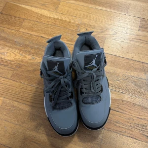 Grå Nike Jordan 4 sneakers - Säljer ett par grå Nike Jordan 4 sneakers med svarta och vita detaljer samt gul insida på plösen. Skorna har snörning, platt sula och rund tå. Ikonisk Jumpman-logga på hälen och plösen. Materialet är skinn och syntet, perfekt för dig som gillar streetwear.