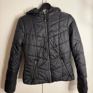 Svart quiltad pufferjacka från Bershka - Svart pufferjacka från Bershka i storlek S. Jackan har huva med snörning, två fickor med dragkedja och ett quiltat mönster. Perfekt för kyliga dagar och har en snygg, modern look med lång ärm och figurnära passform.