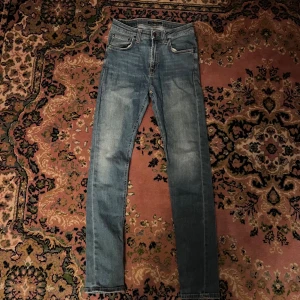 Blå slim jeans från Nudie Jeans 28/30 - Säljer ett par klassiska blå skinny jeans från Nudie Jeans med snygga slitningar och kontrastsömmar. Jeansen har fem fickor, orange broderi på bakfickorna och en brun läderpatch i midjan. Perfekt passform och riktigt sköna att bära. Midja 32cm längd 101cm (52)